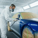 Un carrossier en combinaison et masque, peignant une voiture bleue avec un pistolet dans une cabine de peinture.