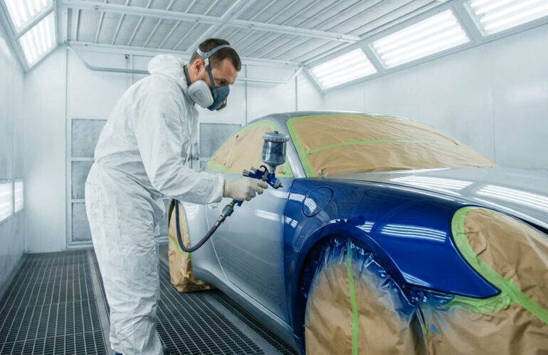 Un carrossier en combinaison et masque, peignant une voiture bleue avec un pistolet dans une cabine de peinture.